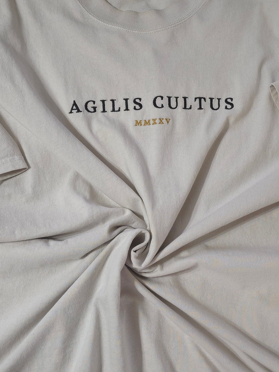 Flat lay of an Agilis Cultus brand cream-colored garment-dyed t-shirt, showing a thick crewneck collar, heavy fabric texture, and a center chest embroidery that reads "AGILIS CULTUS" above a subtle crest.