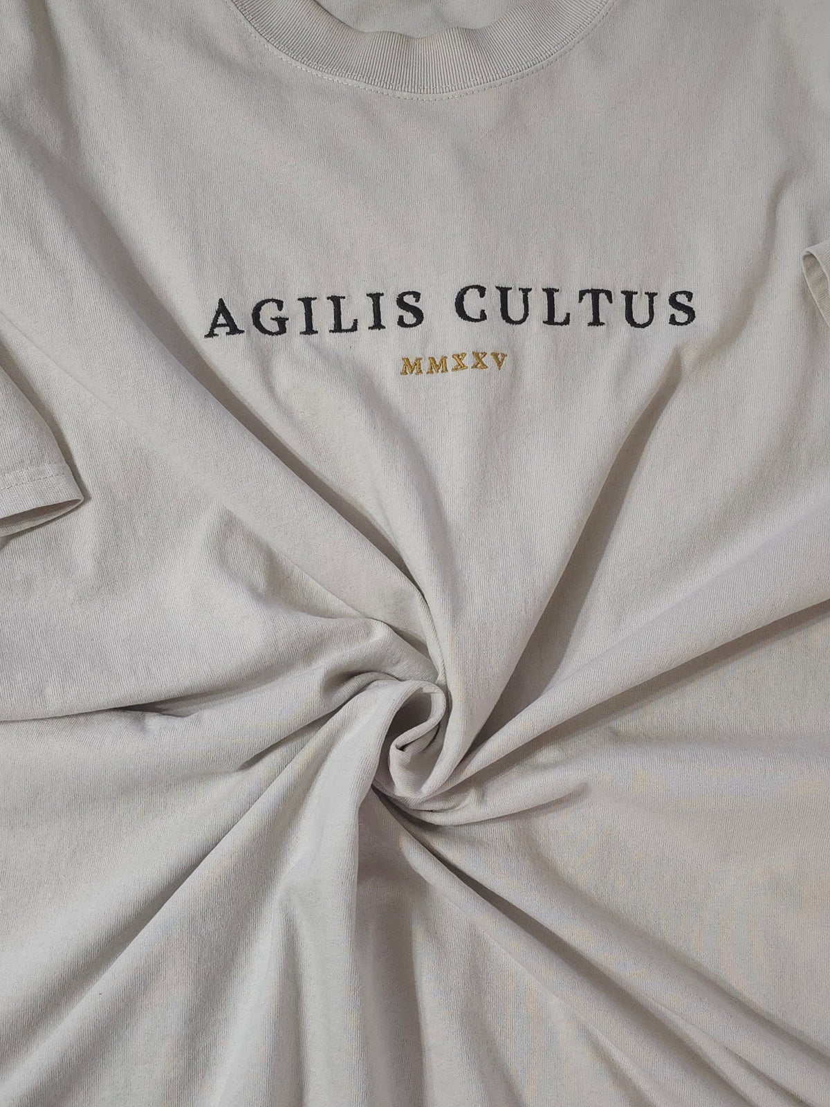 Flat lay of an Agilis Cultus brand cream-colored garment-dyed t-shirt, showing a thick crewneck collar, heavy fabric texture, and a center chest embroidery that reads "AGILIS CULTUS" above a subtle crest.