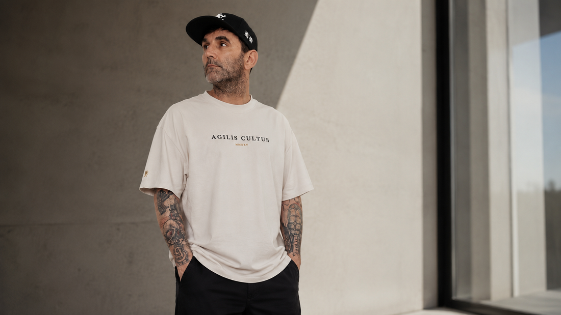 A man wearing a faded bone Agilis Cultus oversized heavyweight t-shirt, black cap, and dark pants stands in a minimalist architectural setting with soft natural light, showing the shirt’s relaxed fit, structured drape, and clean chest branding.